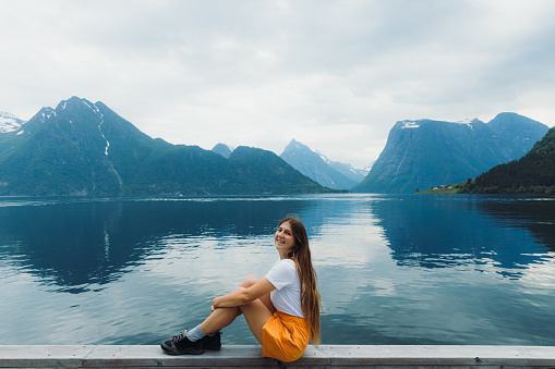 Female traveler with long hair in orange shorts sitting by the seashore admiring the reflecting mountain peaks in Hjorungfjorden, Western Norway