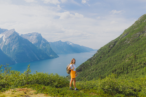 Happy woman in with long hair in orange shorts staying on the top viewpoint with a view of beautiful bay surrounded by snowcapped mountains during summertime in western fjords of Norway