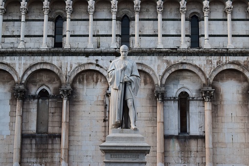 An ornate bronze statue standing atop a white stone pedestal in front of a large, imposing building