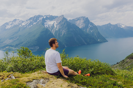 Happy male in white t-shirt sitting on the top viewpoint with a view of beautiful bay surrounded by snowcapped mountains during summertime in western fjords of Norway