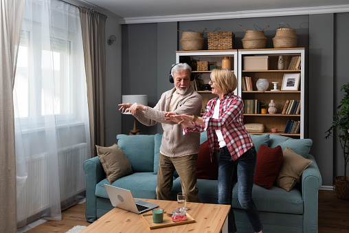 Joyful active old retired romantic couple dancing laughing in living room, happy middle aged wife and elder husband having fun at home, smiling senior family grandparents relaxing bonding together