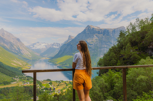 Female traveler with long hair in orange shorts staying on the top admiring a view of beautiful fjord surrounded by green mountain peaks on Western Norway, Scandinavia