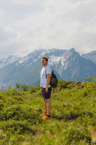 Side view of smiling handsome male with backpack admiring the trip on west fjords of Norway, walking on the top of mountain with idyllic background view of snowcapped mountains