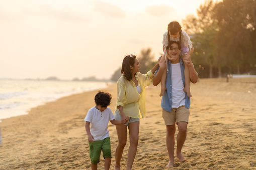 Happy Asian family travel ocean on summer holiday vacation. Parents and children kid enjoy and fun outdoor activity lifestyle walking and playing together on tropical island beach at summer sunset.