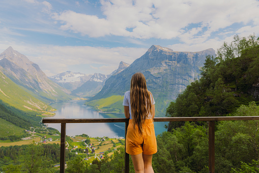 Female traveler with long hair in orange shorts staying on the top admiring a view of beautiful fjord surrounded by green mountain peaks on Western Norway, Scandinavia