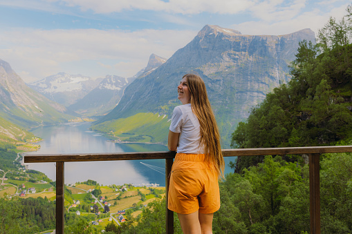 Smiling Female traveler with long hair in orange shorts staying on the top admiring a view of beautiful fjord surrounded by green mountain peaks on Western Norway, Scandinavia