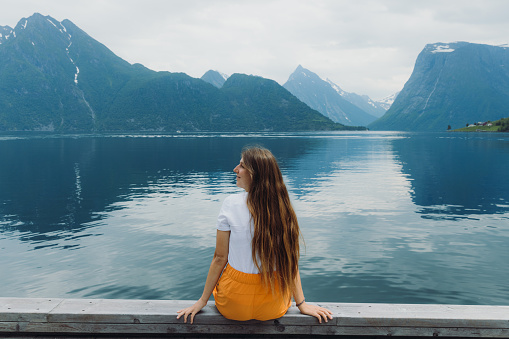 Female traveler with long hair in orange shorts sitting by the seashore admiring the reflecting mountain peaks in Hjorungfjorden, Western Norway