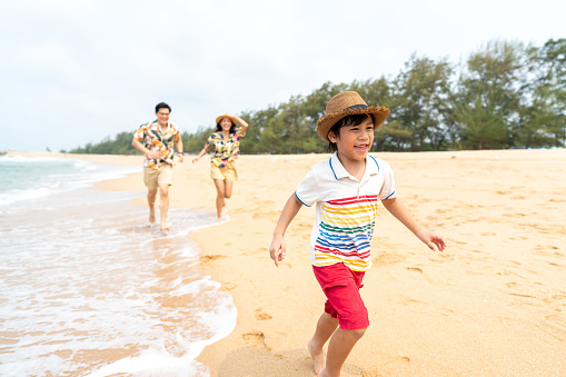 Happy Asian family travel ocean on summer holiday vacation. Parents and little boy son enjoy and fun outdoor activity lifestyle walking and playing together at tropical island beach in sunny day.