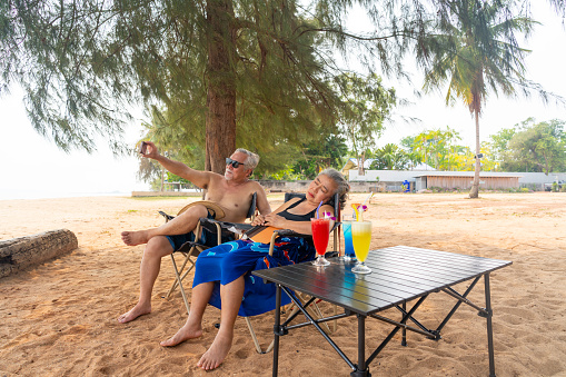 Happy Asian family senior couple in swimwear resting together at tropical beach in sunny day. Retired elderly people enjoy romantic outdoor lifestyle travel nature ocean on summer holiday vacation.