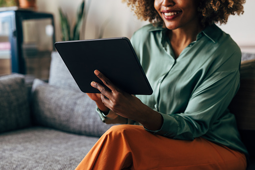 A close up view of an unrecognizable smiling African-American entrepreneur watching something on her tablet while sitting on the sofa in the living room.