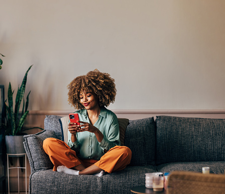 A smiling elegant African-American female using her smartphone while sitting on the cozy sofa in the living room.