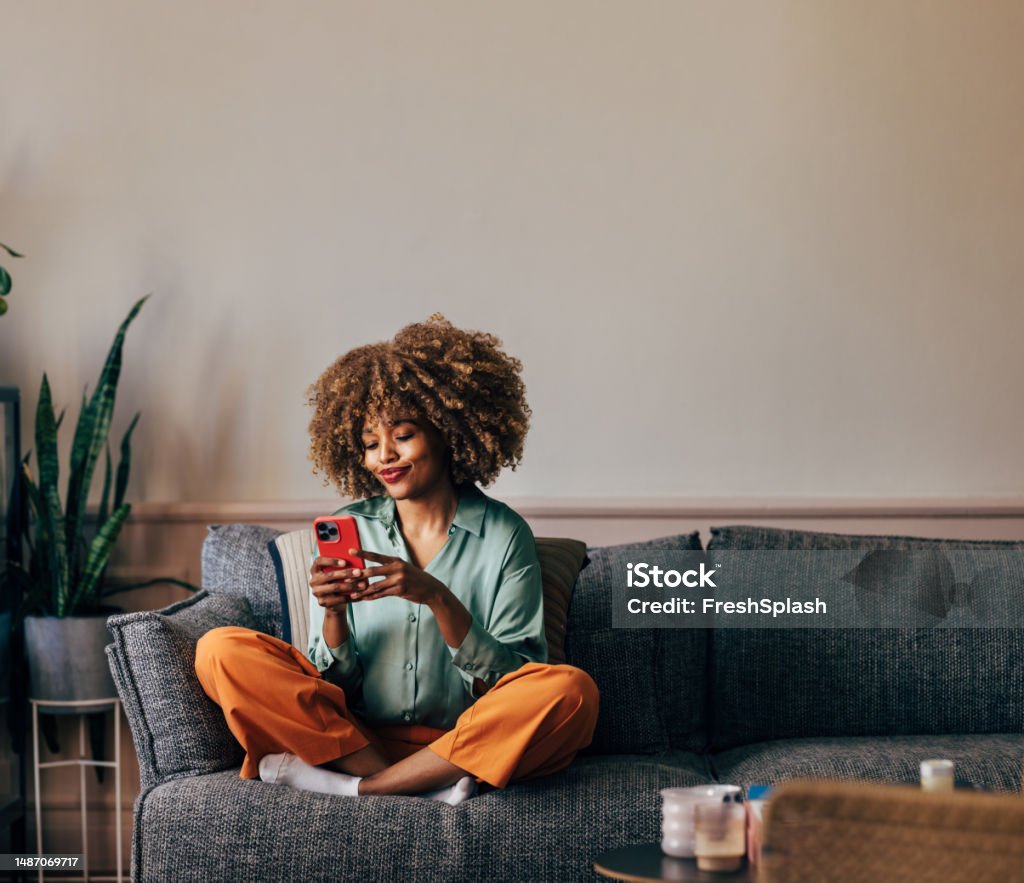 A Happy Beautiful Woman Texting On Her Mobile Phone While Relaxi A smiling elegant African-American female using her smartphone while sitting on the cozy sofa in the living room. Women Stock Photo