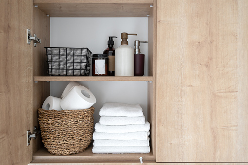 closeup of wooden cabinet with open door and wicker basket with toilet paper, stack of clean folded towels, cosmetics product and bath accessories on shelves inside