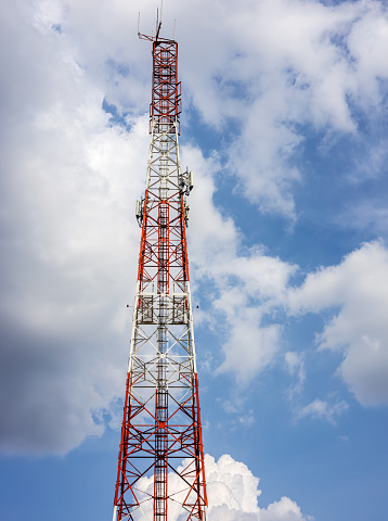 View of a large red-and-white telecommunication tower towering among the clouds in the blue sky as an afternoon backdrop of the Thai countryside.