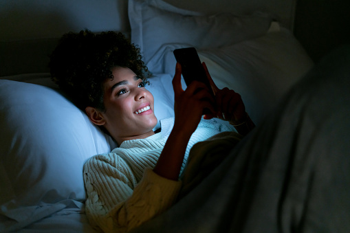 Happy African American young woman lying in bed texting on her cell phone and smiling - lifestyle concepts