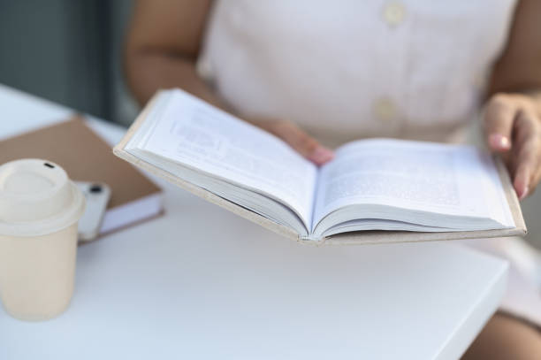 istock Woman with coffee reading book at white table outdoors, closeup 1487081284