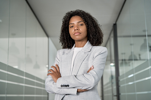 Confident portrait of young African American leader manager, stands proudly, crossed arms and looking at camera in business office center. Portrait of professional business woman in stylish suit.