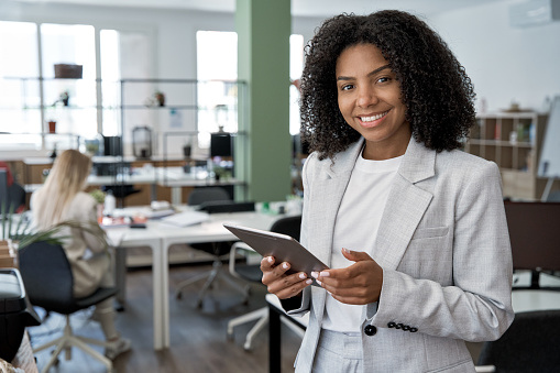 Young business woman using touchscreen pc tablet standing in modern office. African American female businesswoman, successful entrepreneur doing online web work wearing suit and looking at camera.