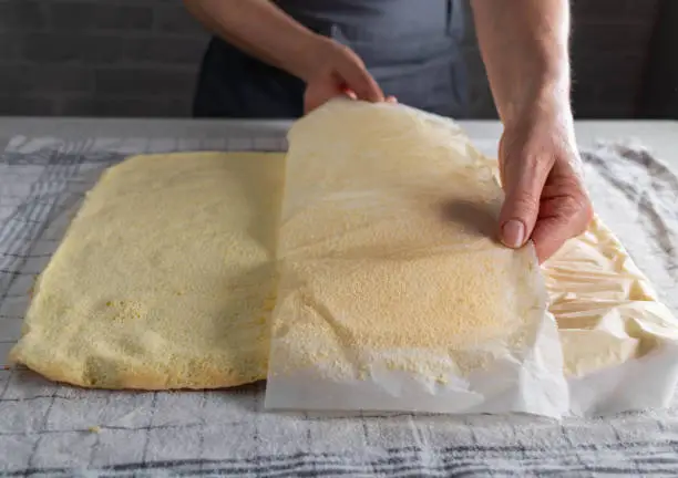 Baking a swiss roll. Removing or pull off baking paper from a fresh baked sponge cake base on kitchen counter by woman´s hands