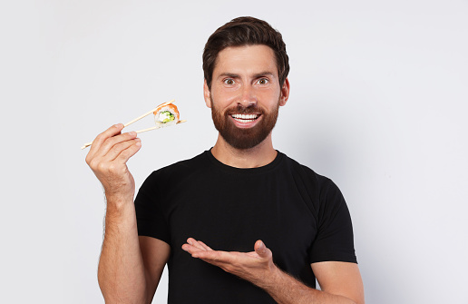 Happy man holding sushi roll with chopsticks on light grey background
