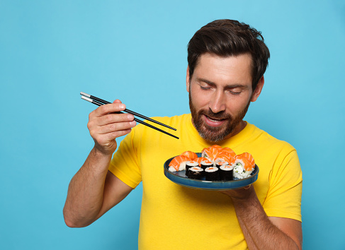 Happy man holding plate with tasty sushi rolls and chopsticks on light blue background
