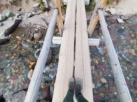 This is a photograph taken on a mobile phone outdoors from a personal point of view of an unrecognizable woman’s boots about to cross a small rustic wooden bridge in Glacier National Park during autumn of 2020.