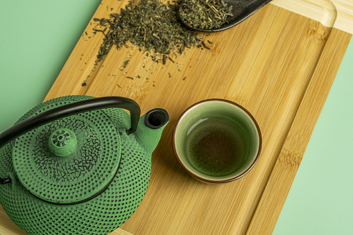 Closeup of green cast iron teapot with green porcelain cup with tea and bamboo wood table
