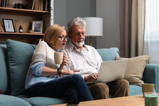 Happy middle aged family couple relaxing on sofa, using computer web surfing information shopping together at home. Elderly man showing laptop apps to retired wife, older generation with tech concept.