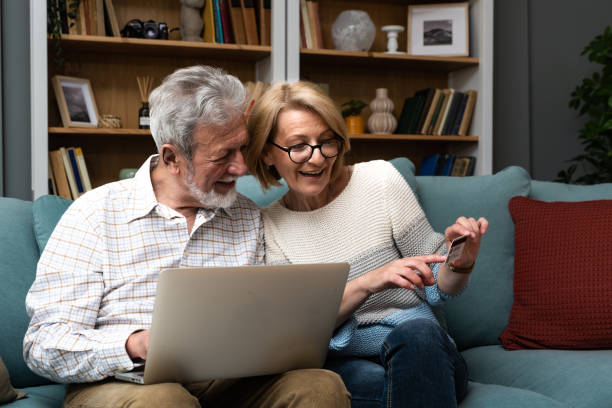 pareja mayor en casa. el apuesto hombre mayor y la atractiva mujer mayor pasan tiempo juntos. sentado en un sofá con computadora portátil y tarjeta de crédito en las manos. compras en línea ordenar o reservar - heterosexual couple love romance couple fotografías e imágenes de stock