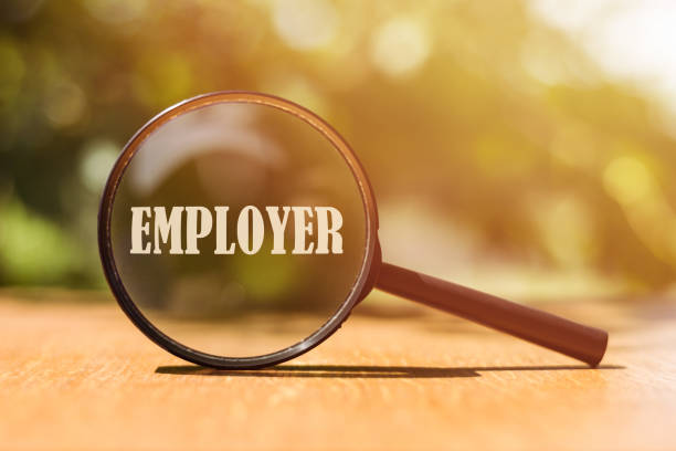 istock Magnifying glass with text Employer on wooden surface over blurred tree background in sunlight. 1487104847