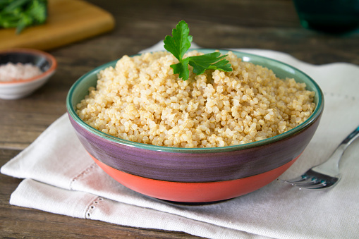 A bowl of cooked quinoa on a dark wooden table with fresh broccolini and Himalayan salt in the background. Healthy vegan food.