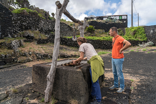 Small picturesque village, houses made of volcanic rocks, at the edge of black lava coast. Two men looking at old stone well in the village, Cabrito, Pico island, Azores