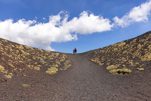 Mount Etna, Sicily, Italy - April 27, 2023: Two people walking the path of the slopes of the volcano Mount Etna on the sunny day. Stones and solidified lava, withered vegetation