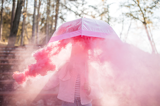 A millennial woman stands under an umbrella in nature, surrounded by pink smoke from a burning torch