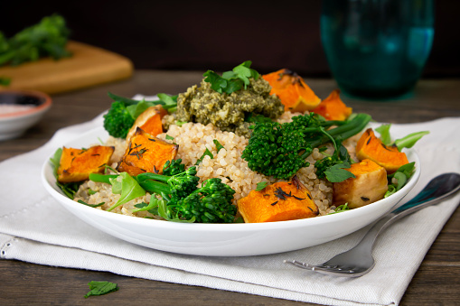 A Quinoa bowl with roasted butternut squash, broccolini, leafy greens and pesto. Shot on a dark wooden table with fresh vegetables and Himalayan salt in the background. \nHealthy vegetarian food recipe.