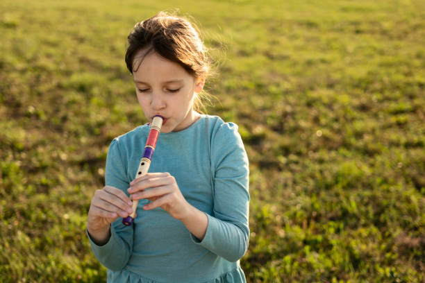 Young girl with long brown hair stands among tall grasses on a sunny autumn day. She wears a blue plaid shirt and examines something in her hands with a curious, happy expression. Sunlight filters through the foliage, creating a warm, natural atmosphere in the outdoor setting.