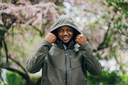 A handsome black smiling man puts a hood on his head while standing in a park and looking directly at the camera