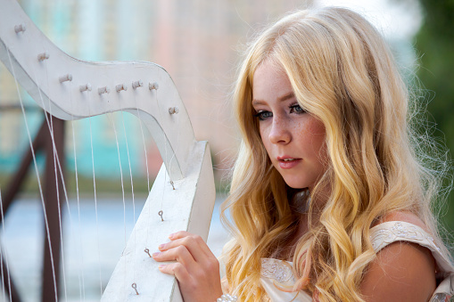 Close-up portrait of beautiful preteen girl with curly blond hair and facial freckles wearing white dress and playing outdoor on harp. Selective focus. Copy space for your text. Music theme.