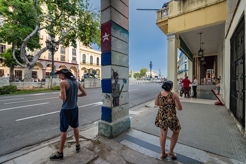 Havana, Cuba – June 21, 2018: Paseo del Prado famous place in Havana with young girl walking and a young boy standing. A column with Cuban flag painted in a blue sky day.