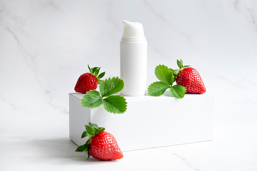 Blank white plastic skincare tube w/ fresh strawberries fruit on pink background. Natural cosmetic beauty product branding mock-up for brightening cream, body lotion, facial foam or shampoo. Flat lay.