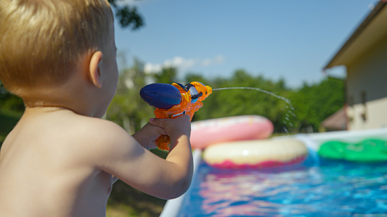 CLOSE UP: Rear view shot of a little boy spraying with water gun while water fight. Playful family holiday moment of a child at garden pool. Fun and refreshing outdoor activities for hot summer days.