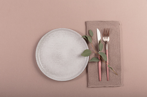 Festive table setting. Silver modern cutlery, eucalyptus branch and porcelain plate as mock up on beige table background. Mediterranean wedding or restaurant menu concept. Flat lay, top view