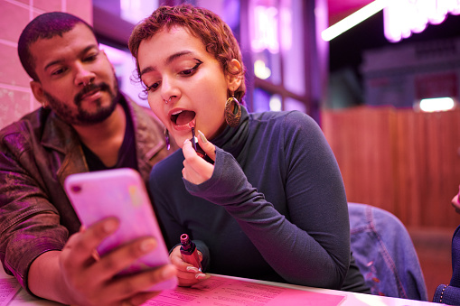 Man using a phone screen to help his friend put on lipstick while sitting at a table in a bar during night out