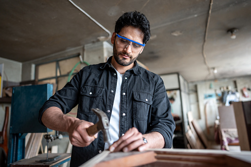 Mid adult carpenter hammering a nail on piece of wood at carpentry