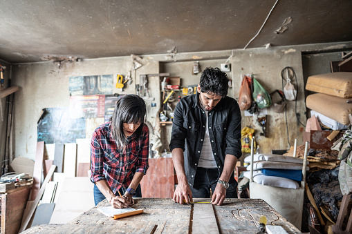 Mid adult man taking measurements a piece of wood while his friend takes notes on a notepad at carpentry