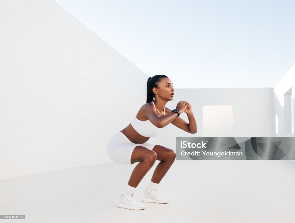 Female fitness influencer doing sit-ups in a white outdoor studio. Young woman doing workout outdoors. Female in white fitness attire is doing exercises for her glutes. Exercising Stock Photo