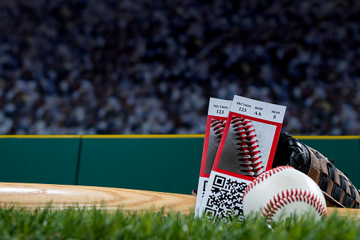 A low angle view of a pair of ticket stubs resting against a baseball bat, glove and baseball sitting on a synthetic grass turf with green padded wall around the sports field during a night game.