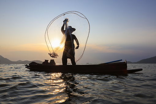 Fisherman casting his net on during sunrise.Silhouette Asian fisherman on wooden boat casting a net for freshwater fish,Silhouette Fisherman Fishing Nets on the boat.Thailand