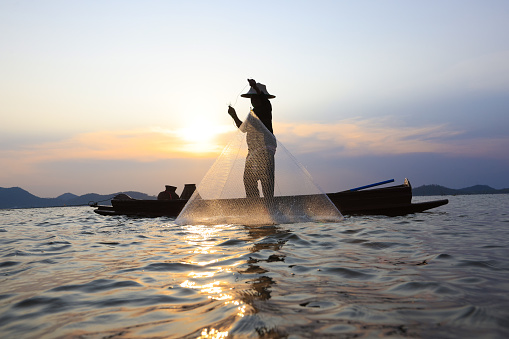 Fisherman casting his net on during sunrise.Silhouette Asian fisherman on wooden boat casting a net for freshwater fish,Silhouette Fisherman Fishing Nets on the boat.Thailand