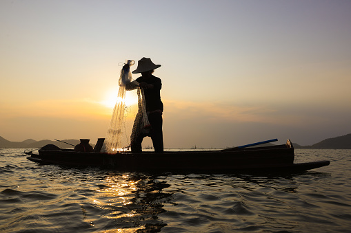 Fisherman casting his net on during sunrise.Silhouette Asian fisherman on wooden boat casting a net for freshwater fish,Silhouette Fisherman Fishing Nets on the boat.Thailand